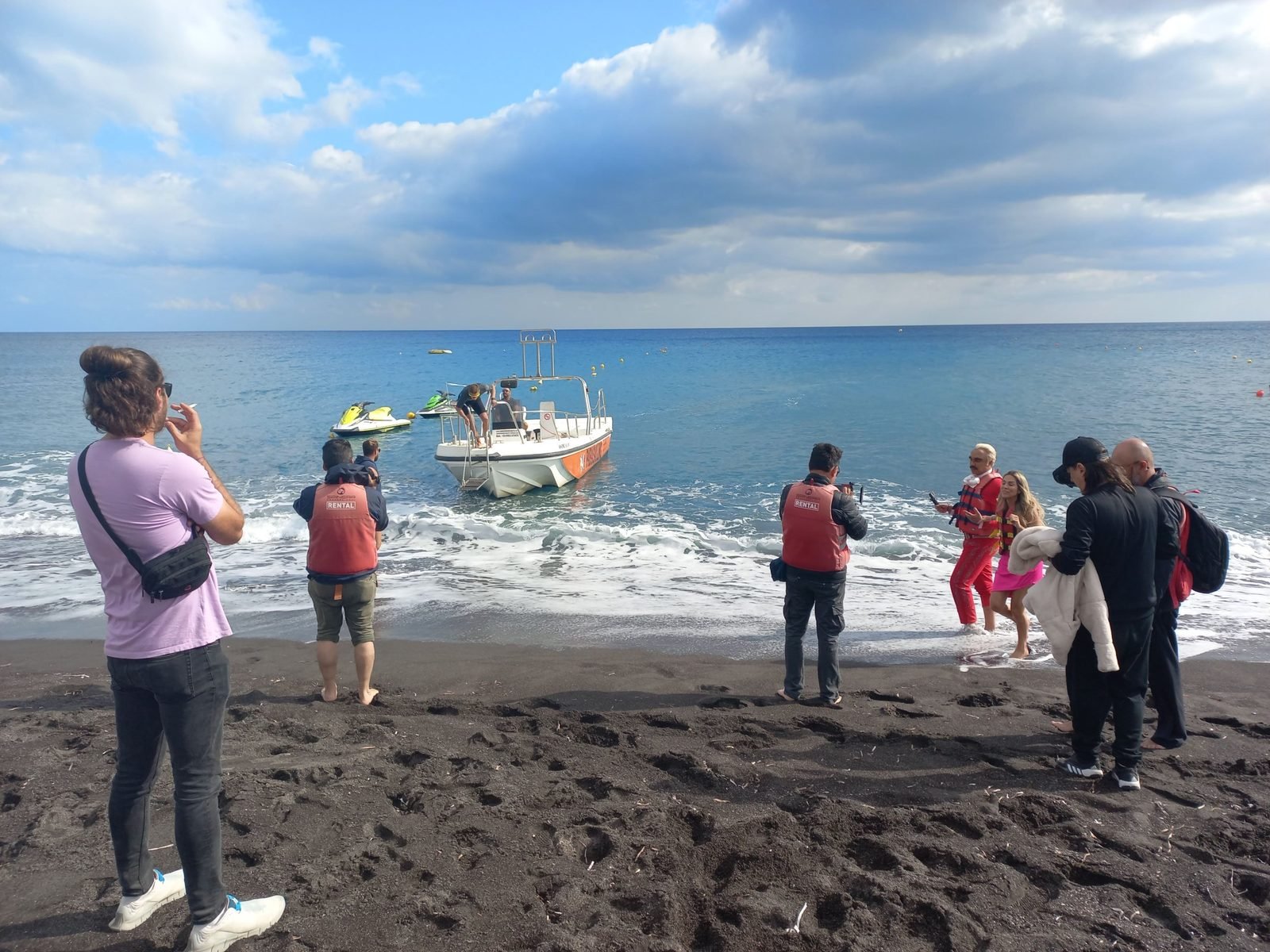 The crew filming the presenters in life jackets on Perivolos black-sand beach, with the rental boat and jet skis, October 2022